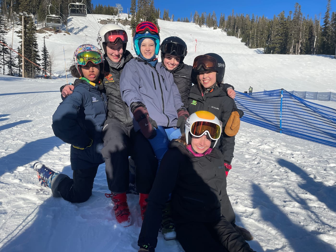 Six teammates piled on top of each other laughing on a bluebird day, the race hill behind them